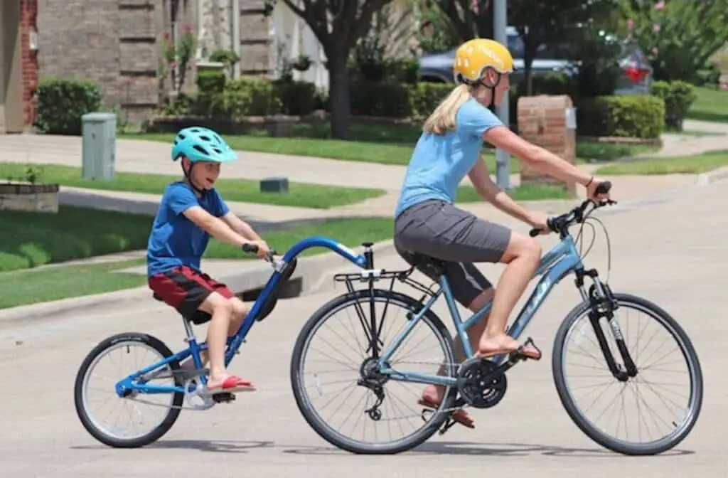 tandem biling shows an adult in the front seat and a child on a smaller bike attached to the adult bike. Both are wearing helmets.